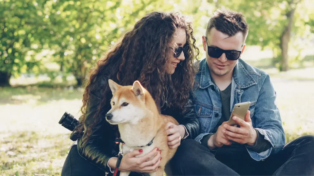 Couple sitting in a park with a Shiba Inu dog, both looking at a smartphone together.